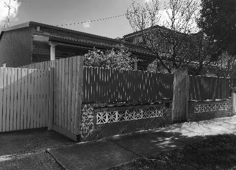 Black and white photo of a house with a fenced yard and a tree.